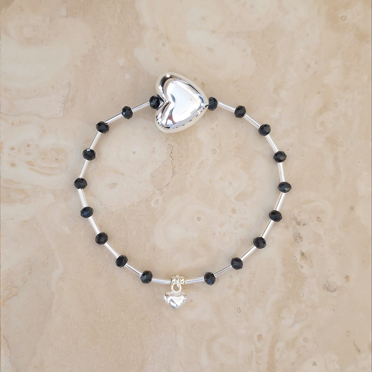 A beaded stretch bracelet featuring alternating black and silver beads, a large polished silver heart charm at the top, and a small silver heart pendant at the bottom, displayed on a beige stone surface.